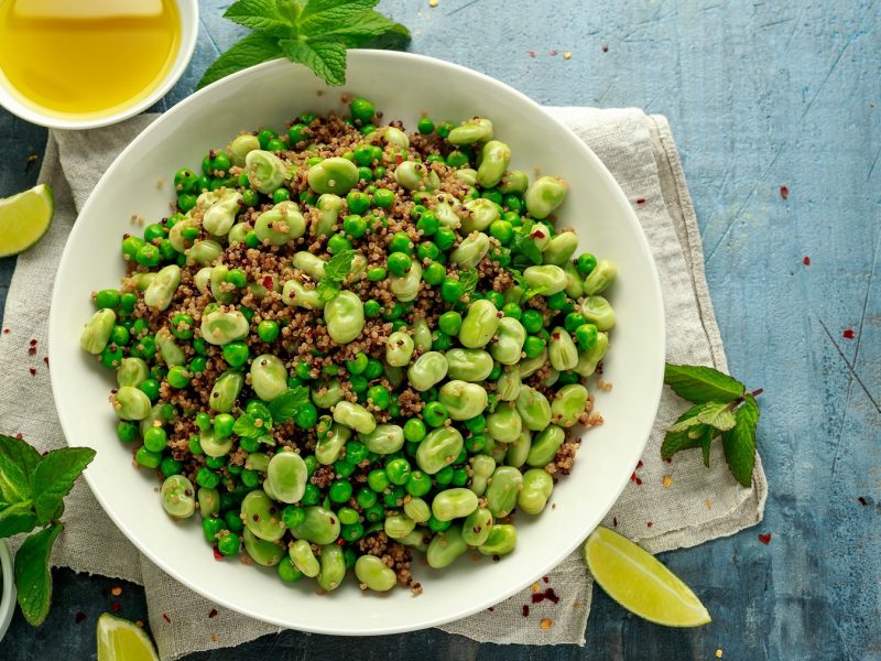 Saubohnensalat mit Quinoa und Erbsen in einer weißen Schüssel, daneben frische Minze, eine Schale mit Öl und Limetten.