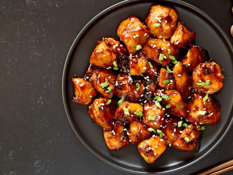 Ein schwarzer Teller mit dem Teriyaki-Hähnchen auf dunklem Untergrund. Rechts neben dem Teller diverse Zutaten für die Sauce.