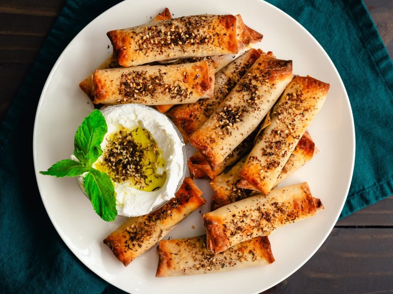 Za'atar-Halloumi-Röllchen mit einer Schale Labneh mit Olivenöl und Za'atar auf einem weißen Teller vor dunklem Hintergrund.