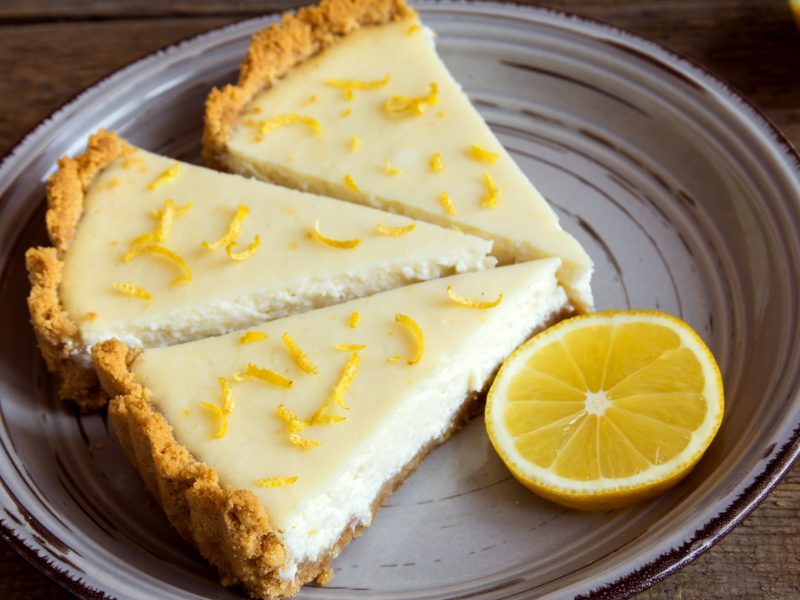 Ein grauer Teller mit drei Stücken vom Zitronen-Cheesecake auf einem rustikalen Holztisch. Alles von schräg oben fotografiert.