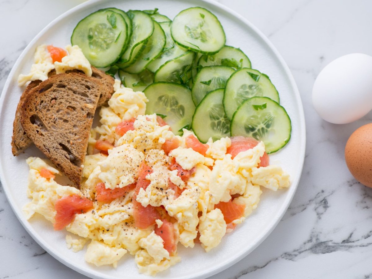 Rührei mit Lachs auf einem weißen Teller serviert mit einer Scheibe Sauerteigbrot und Gurkensalat. Der Hintergrund ist weiß und drei Eier sind darauf verteilt.
