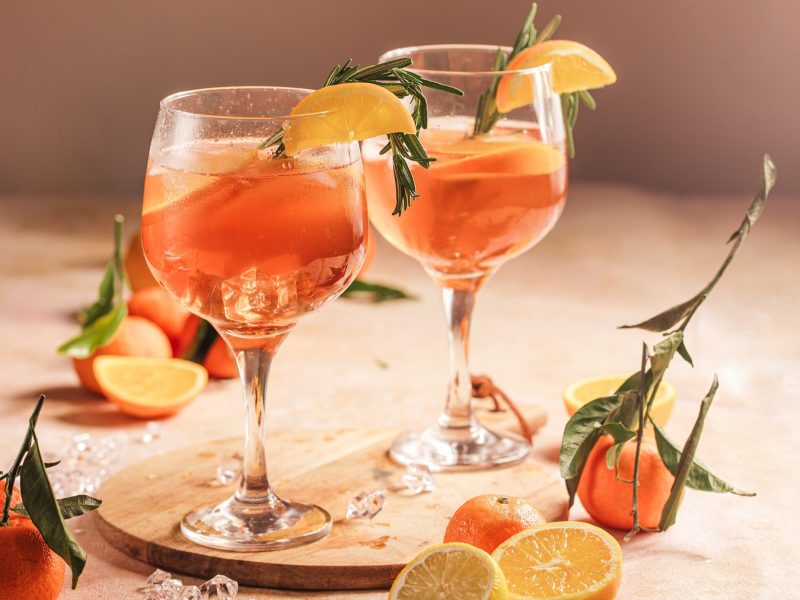 Zwei Aperol Lemon Spritz auf einem hellen Tisch mit Zitronen und Orange. Alles von der Seite fotografiert.