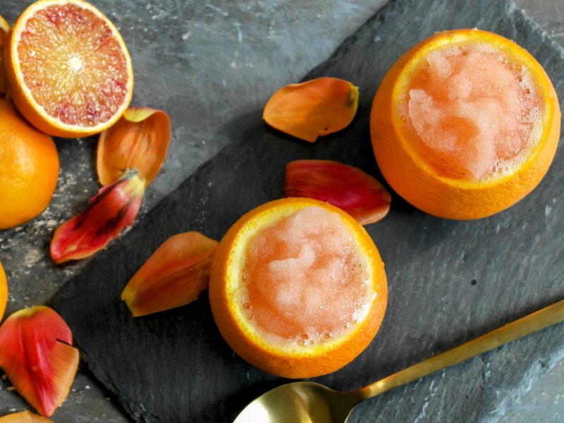 Zwei Orangen mit dem Eis auf einer schwarzen Schiefertafel und grauem Tisch. Ringsherum halbierte Orangen und rötliche Blütenblätter. Alles direkt von oben fotografiert.