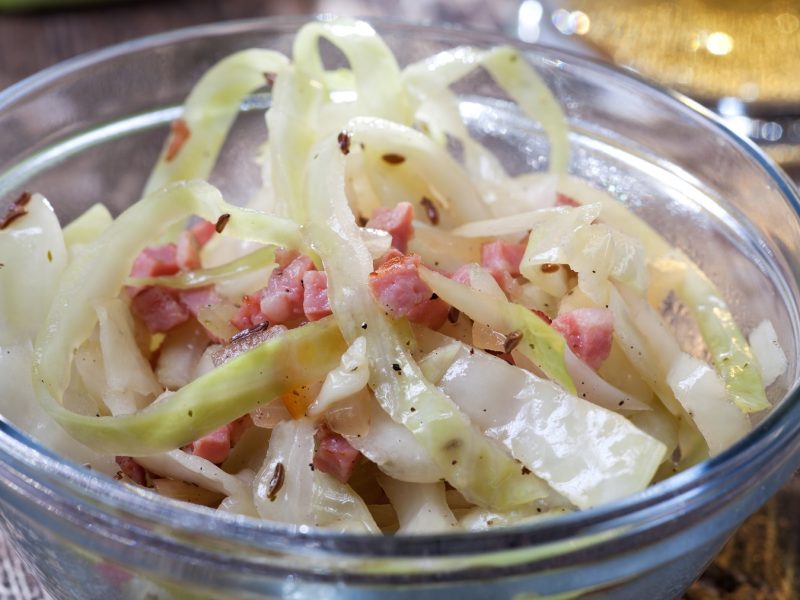 Ein sehr nahes Foto einer kleinen Glasschüssel mit dem Krautsalat auf einem rustikalen Holztisch von leicht schräg oben fotografiert. Im Hintergrund noch unscharf etwas Öl und Weißkohl erkennbar.