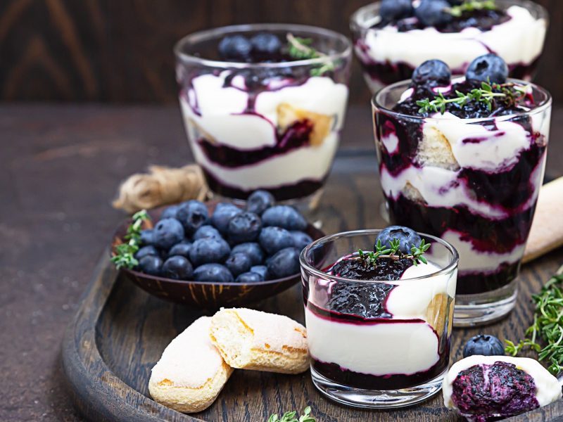 Vier Blaubeer-Mascarpone-Trifle in Gläser vor dunklem Hintergrund.