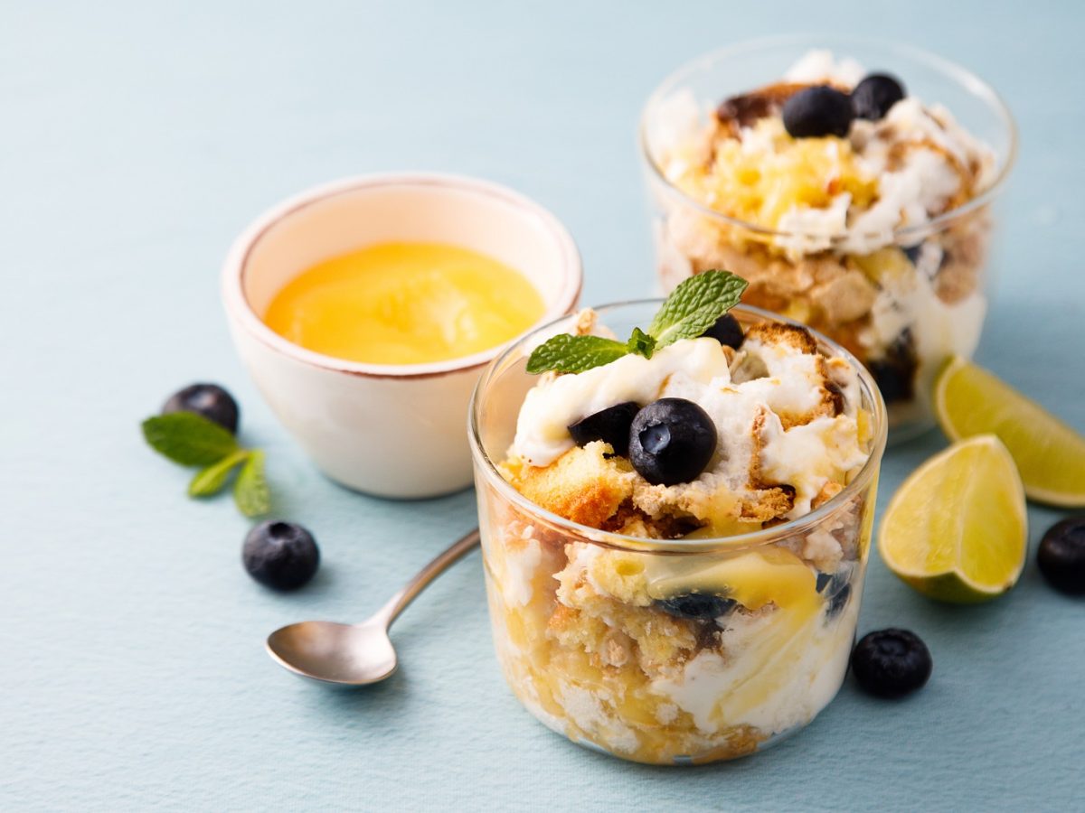 Heidelbeer-Zitronen-Trifle in zwei Gläsern vor blauem Hintergrund, daneben ein Schälchen mit Lemon Curd.