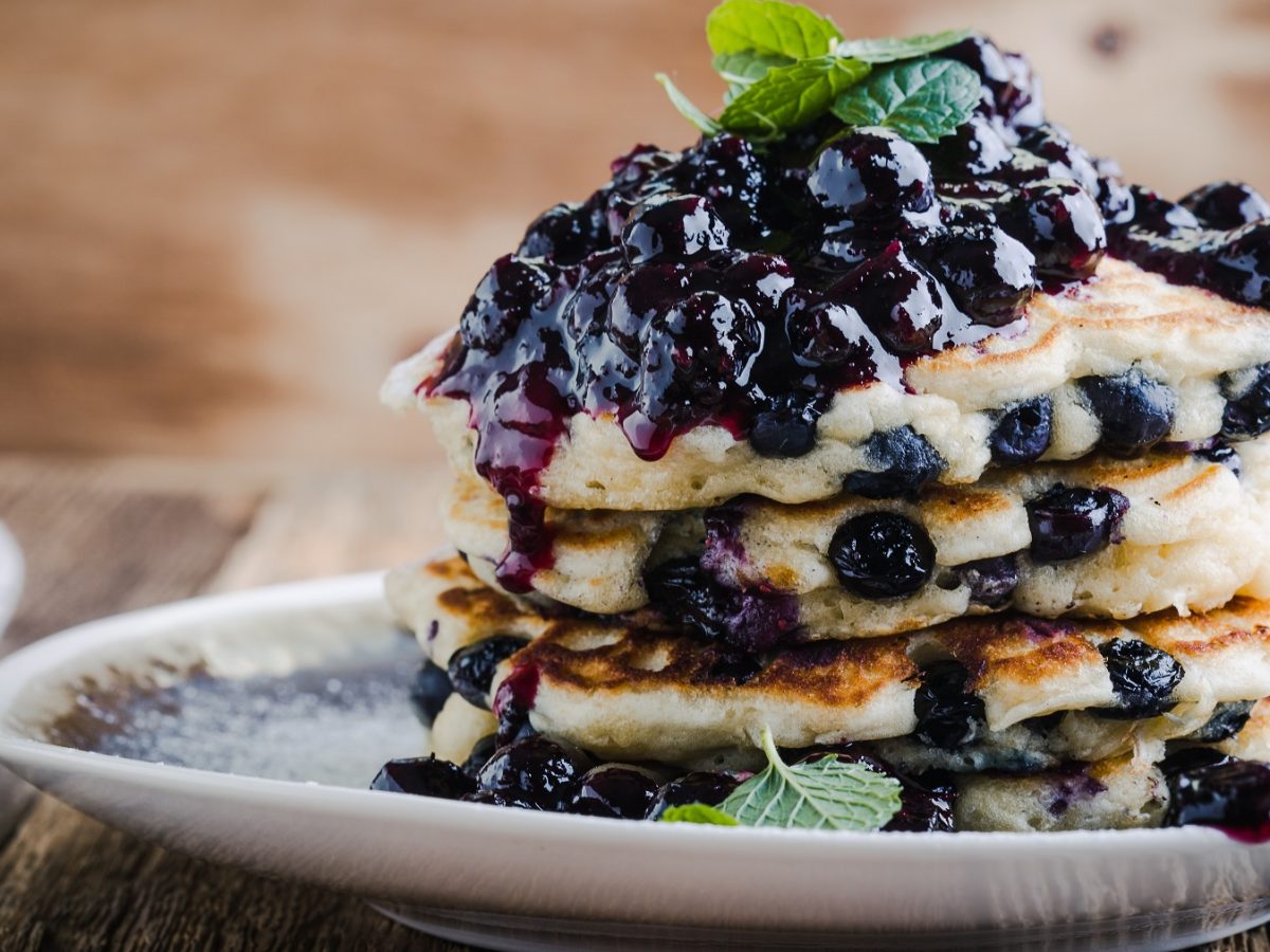 Ein Stapel Blaubeer Pancakes mit reichlich Blaubeerkompott und Minzblättern auf einem Teller vor einem rustikalen Hintergrund.