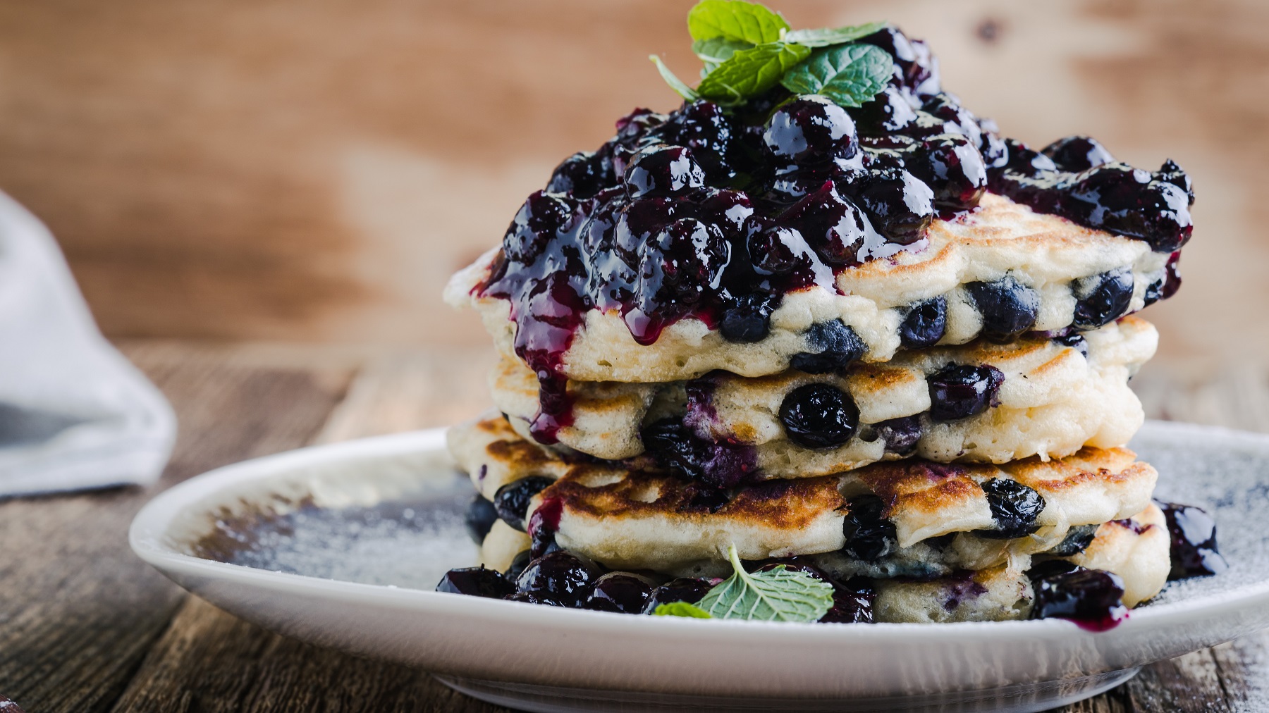 Ein Stapel Blaubeer Pancakes mit reichlich Blaubeerkompott und Minzblättern auf einem Teller vor einem rustikalen Hintergrund.