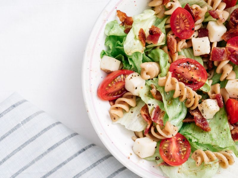 Frischer Nudelsalat mit Tomaten, Salatblättern, Käsewürfeln und Speck auf weißem Hintergrund, daneben ein gestreiftes Tuch.