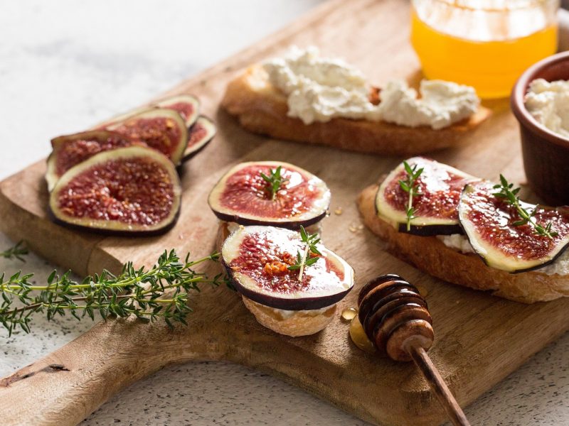 Bruschette mit Ziegenkäse, Honig und Feigen auf einem Holzbrett, davor Honiglöffel.