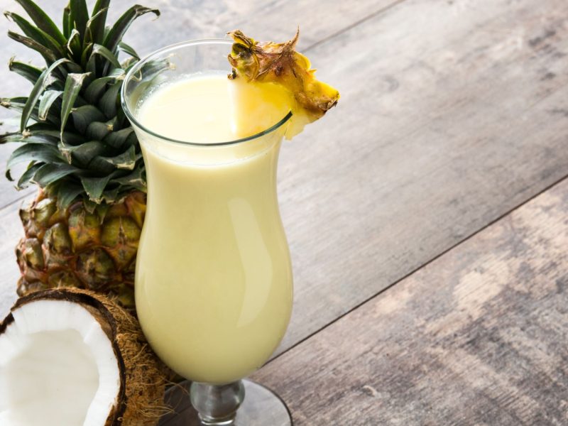 Erfrischender Coconut Kiss Cocktaill mit Ananasscheibe in einem hohen Glas, umgeben von einer Ananas und einer halben Kokosnuss.