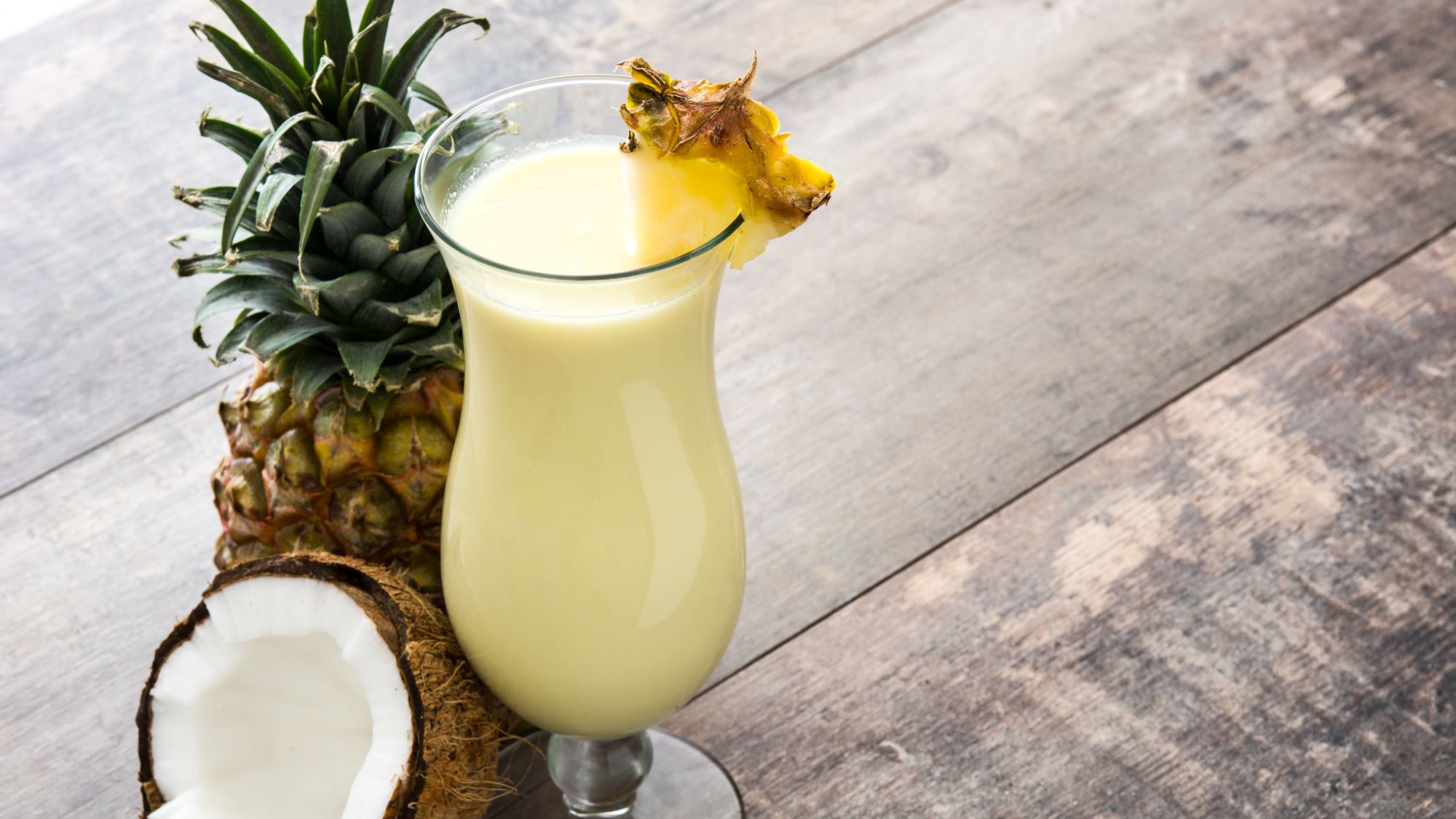 Erfrischender Coconut Kiss Cocktaill mit Ananasscheibe in einem hohen Glas, umgeben von einer Ananas und einer halben Kokosnuss.