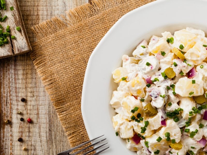 Ein weißer Teller mit dem DDR-Kartoffelsalat direkt von oben auf rustikalem Holztisch, mit Holzbrett fotografiert. Oben links im Bild frischer Schnittlauch, Zwiebel und Pfeffer.