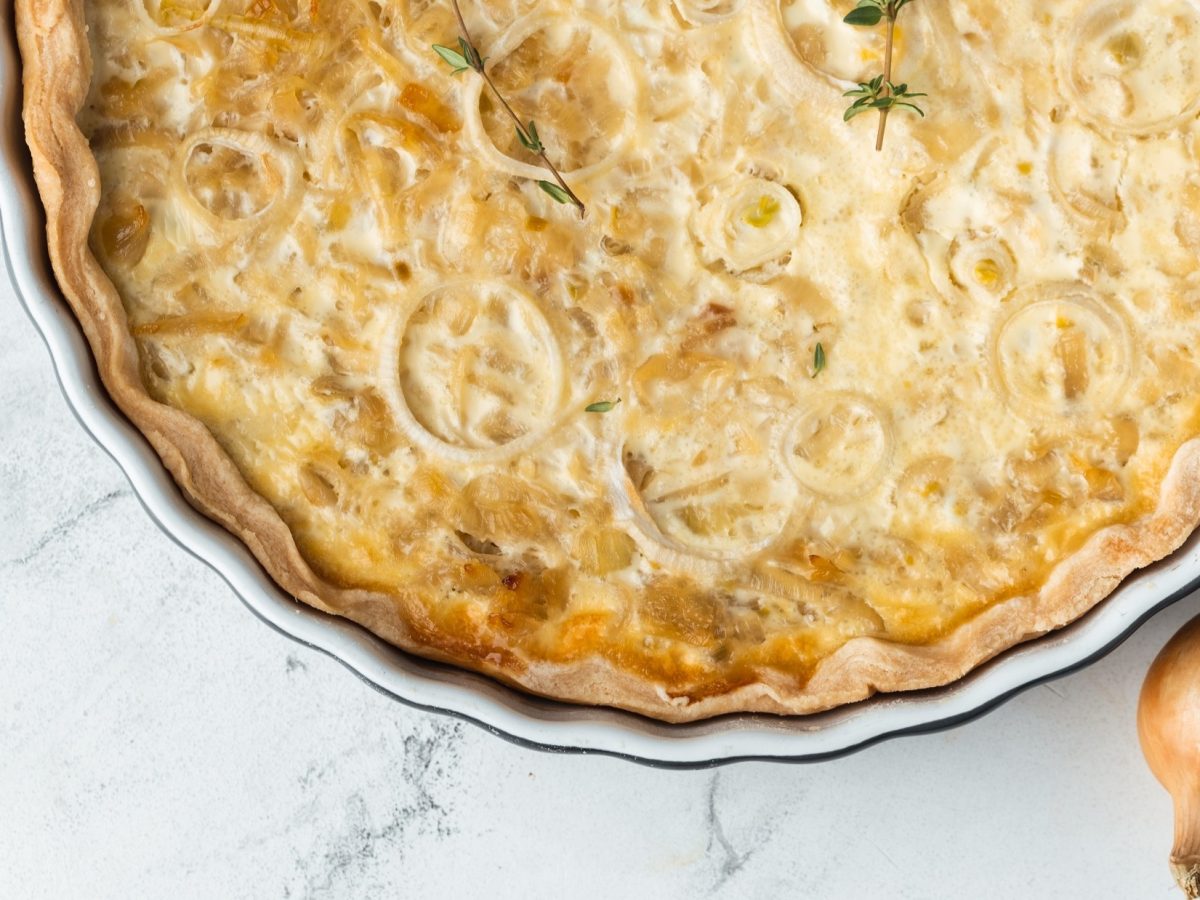 Draufsicht: Auf hellem Marmorboden ist die Hälfte einer runden Form mit einem Elsässer Zwiebelkuchen zu sehen.