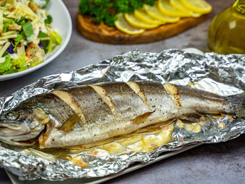 Draufsicht, schräg: Eine ganze gegarte Forelle aus dem Backofen liegt auf buttrigem Alufolie. Im Hintergrund ist eine Schüssel mit Salat sowie einige Zitronenscheiben.