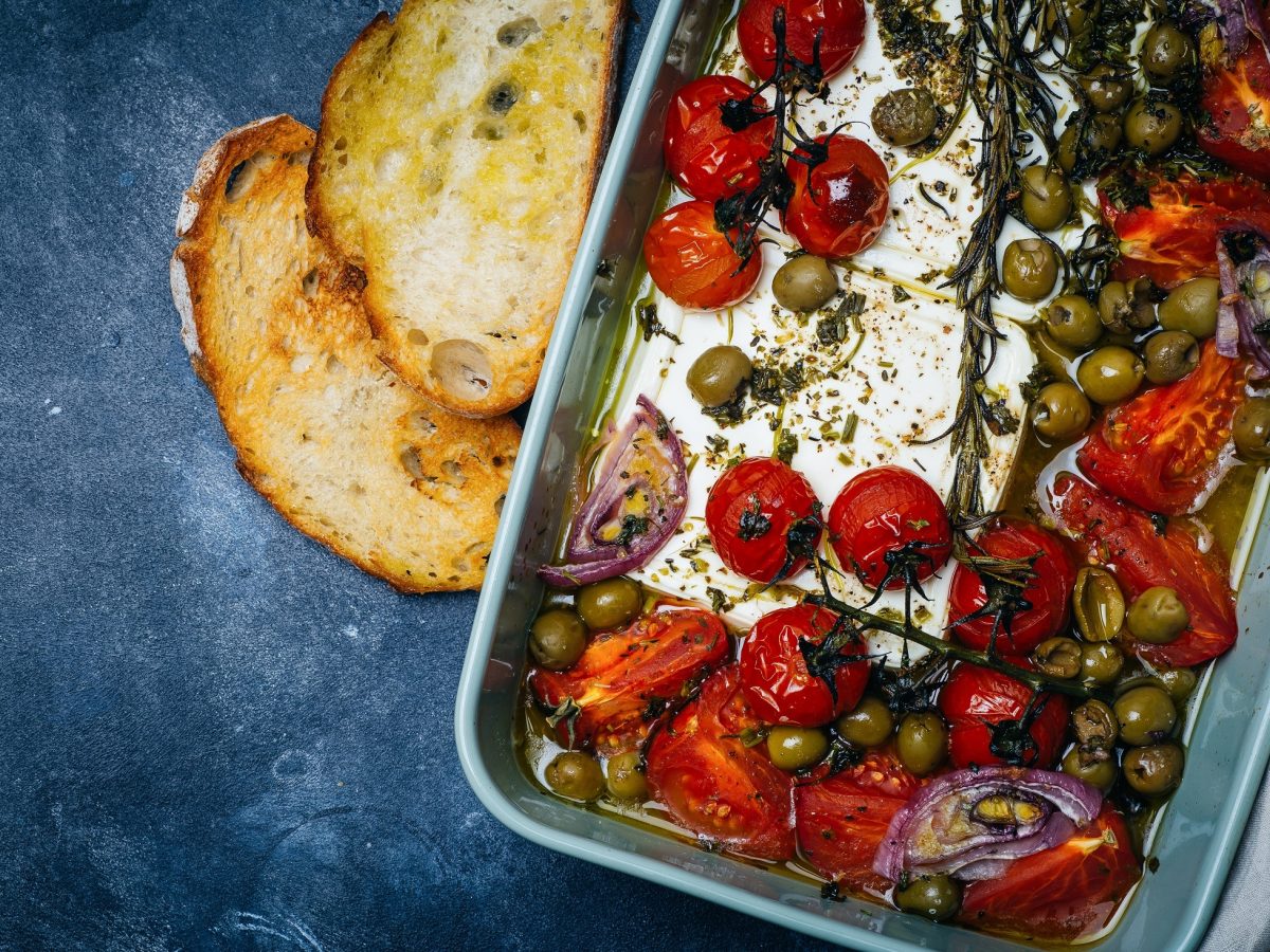 Draufsicht: Auf blauem Untergrund ist eine Auflaufform mit gebackenes Gemüse mit Feta. Es handelt sich um Tomaten, rote Zwiebeln, Oliven und Kräutern.