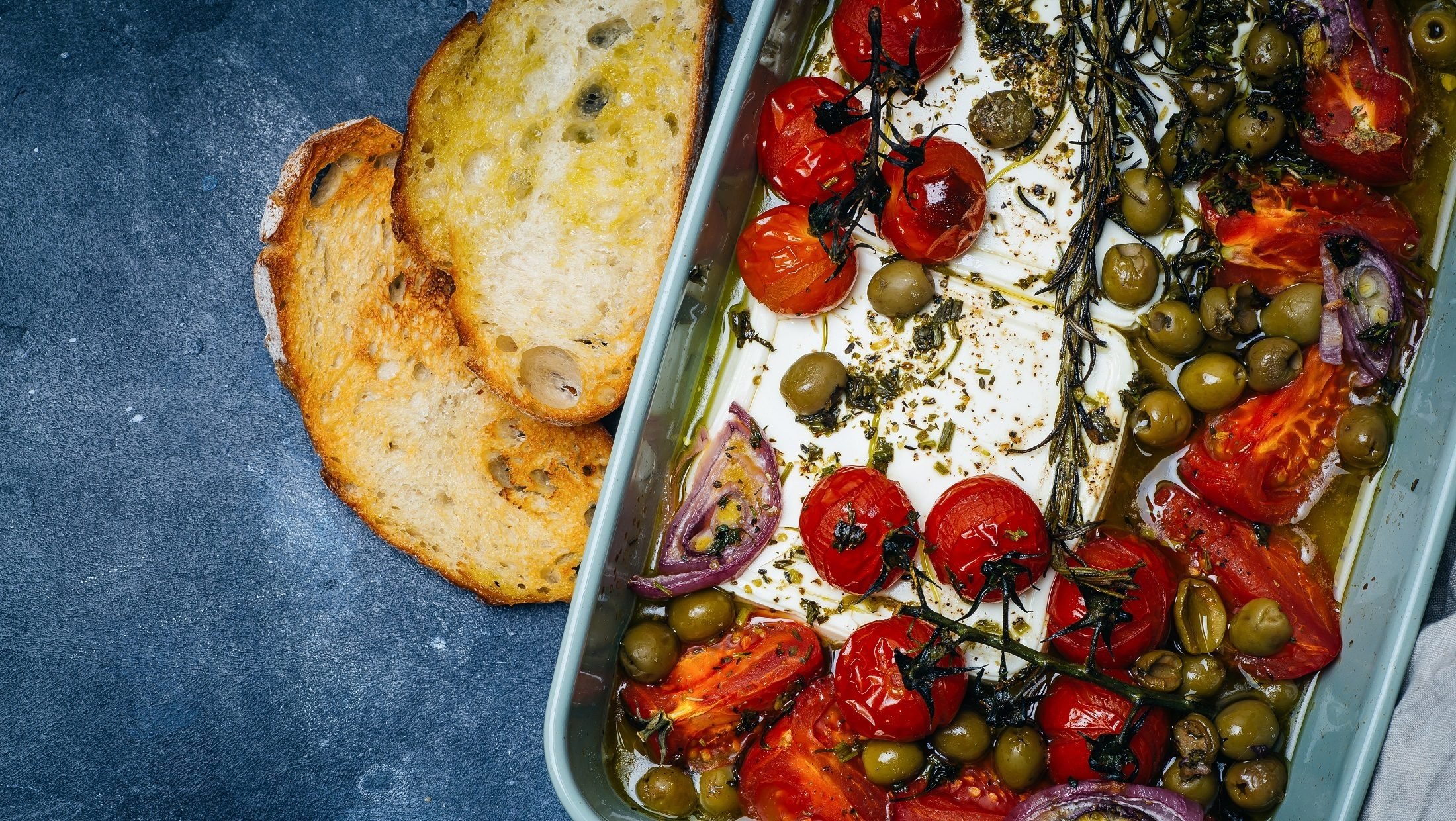 Draufsicht: Auf blauem Untergrund ist eine Auflaufform mit gebackenes Gemüse mit Feta. Es handelt sich um Tomaten, rote Zwiebeln, Oliven und Kräutern.