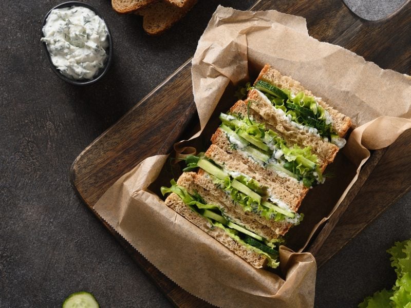 Draufsicht: Fünf verpackte Gurken-Sandwich mit Käse in einer offenen Schachtel, die mit Papier ausgelegt ist. Die Kiste steht auf einem schwarzem Untergrund.