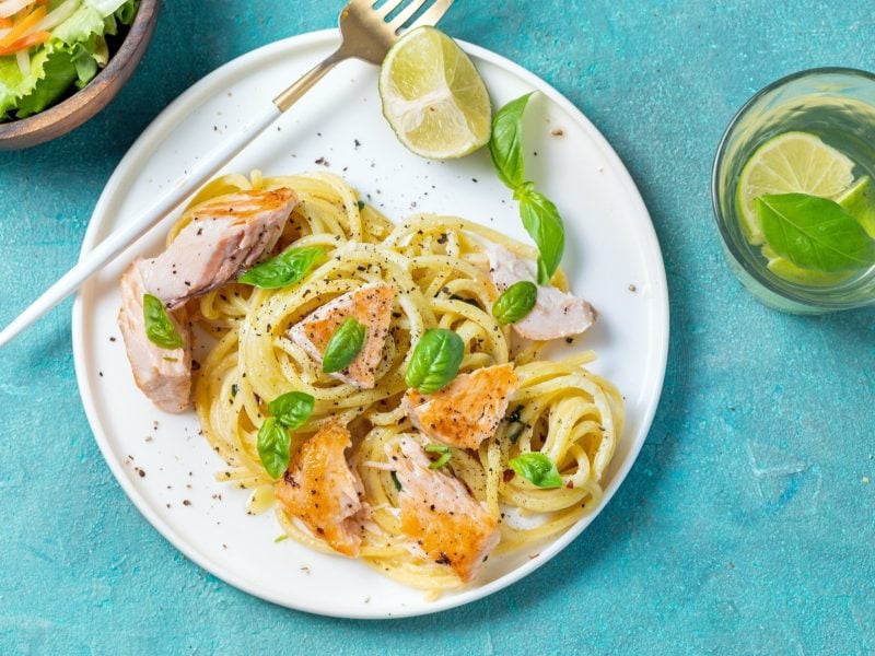 Ein weißer Teller Lachs-Nudeln in Limettensauce auf einem leicht türkises, blauem Untergrund. Daneben sind eine kleine Schüssel mit Salat und ein Glas Wasser mit einer Limette.