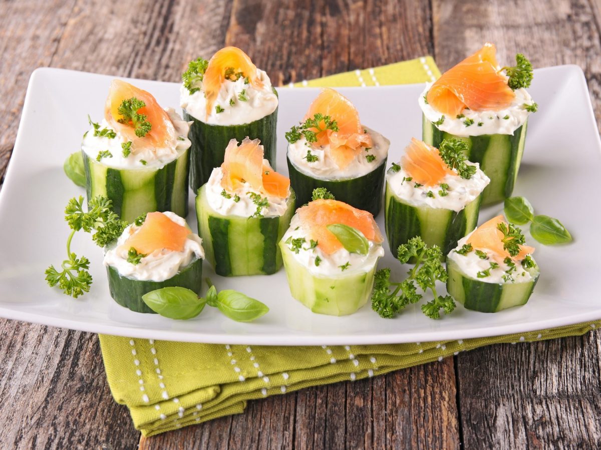 Frontal: Mehrere mit Lachs gefüllte Salatgurken stehen auf einem weißen Teller. Unter der Fingerfood-Platte liegt ein grünes Tuch mit einem weißen Muster. Und das liegt auf einem Boden aus groben Holzplanken.