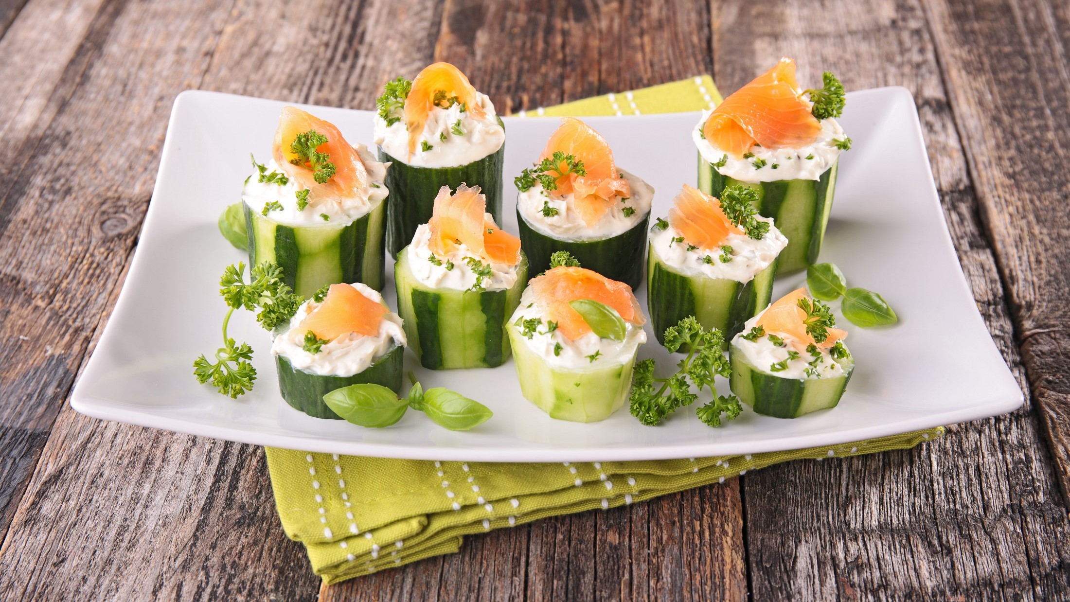 Frontal: Mehrere mit Lachs gefüllte Salatgurken stehen auf einem weißen Teller. Unter der Fingerfood-Platte liegt ein grünes Tuch mit einem weißen Muster. Und das liegt auf einem Boden aus groben Holzplanken.
