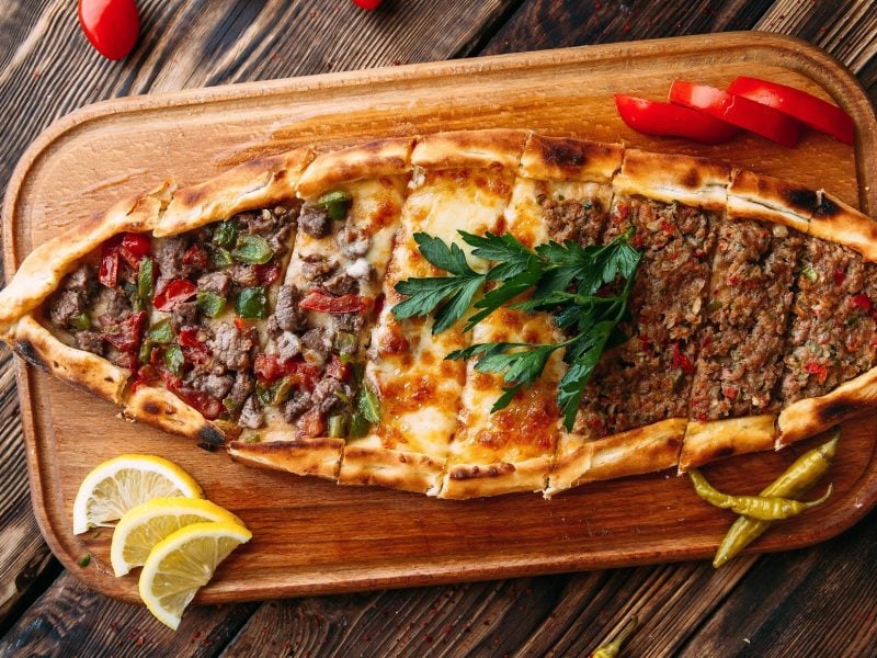 Draufsicht: Ein Holzbrett mit einer selbst gemachten türkischen Pide mit Hackfleisch. Das liegt auf einem Holzboden.