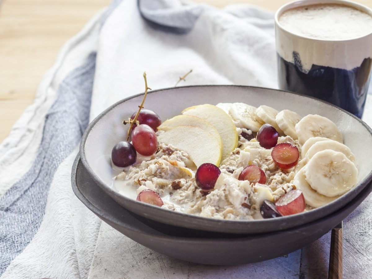 Draufsicht, schräg: Eine rustikale Schale Porridge mit Trauben, Äpfeln und Banane steht auf einem weißen Küchenhandtuch, das blaue Streifen am Rand hat.
