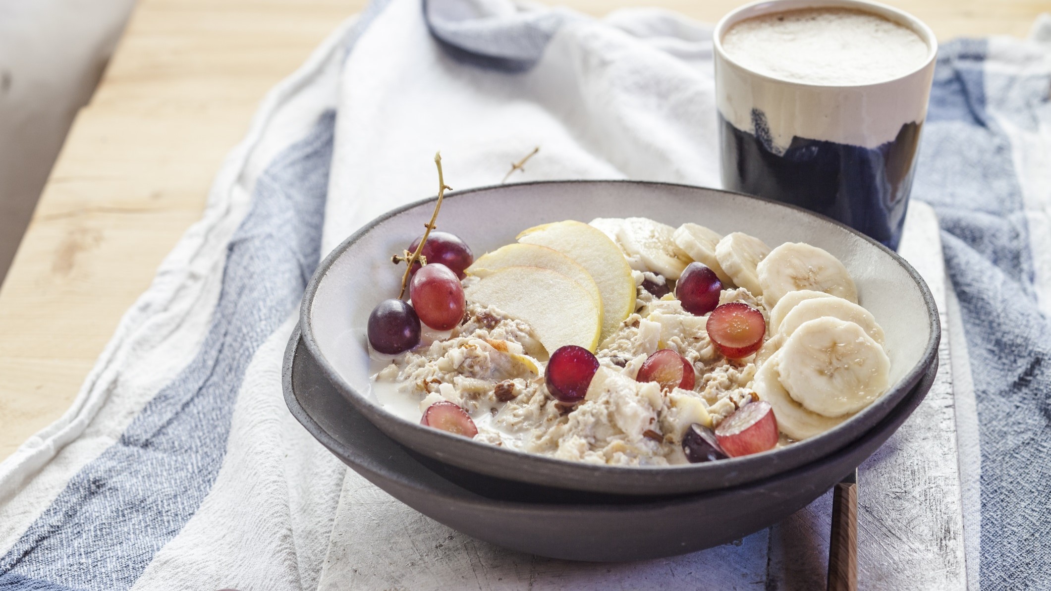 Draufsicht, schräg: Eine rustikale Schale Porridge mit Trauben, Äpfeln und Banane steht auf einem weißen Küchenhandtuch, das blaue Streifen am Rand hat.