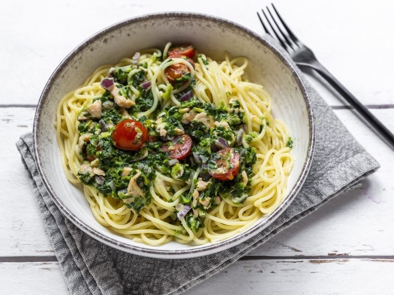 Draufsicht: Eine runde weiße Schüssel mit Spaghetti in Spinat-Lachs-Sauce auf einem weißen Untergrund aus Holz. Die Schüssel steht auf einem grauen Tuch. Daneben liegt eine Gabel.