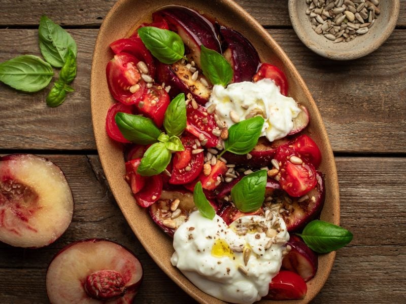 Draufsicht: Ein Tomaten-Pfirsich-Salat mit Burrata in einer ovalen Form. Die steht auf einem Untergrund aus rustikalen Holzplanken. Daneben liegen Pfirsich-Hälften und Basilikumblätter.