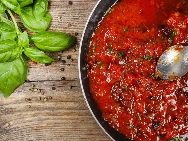 Draufsicht: eine Pfanne mit Tomatensauce, die nach einem Basis-Rezept zubereitet wurde. Die Pfanne steht auf einem Untergrund aus Holz. Daneben liegen frische Basilikumblätter.