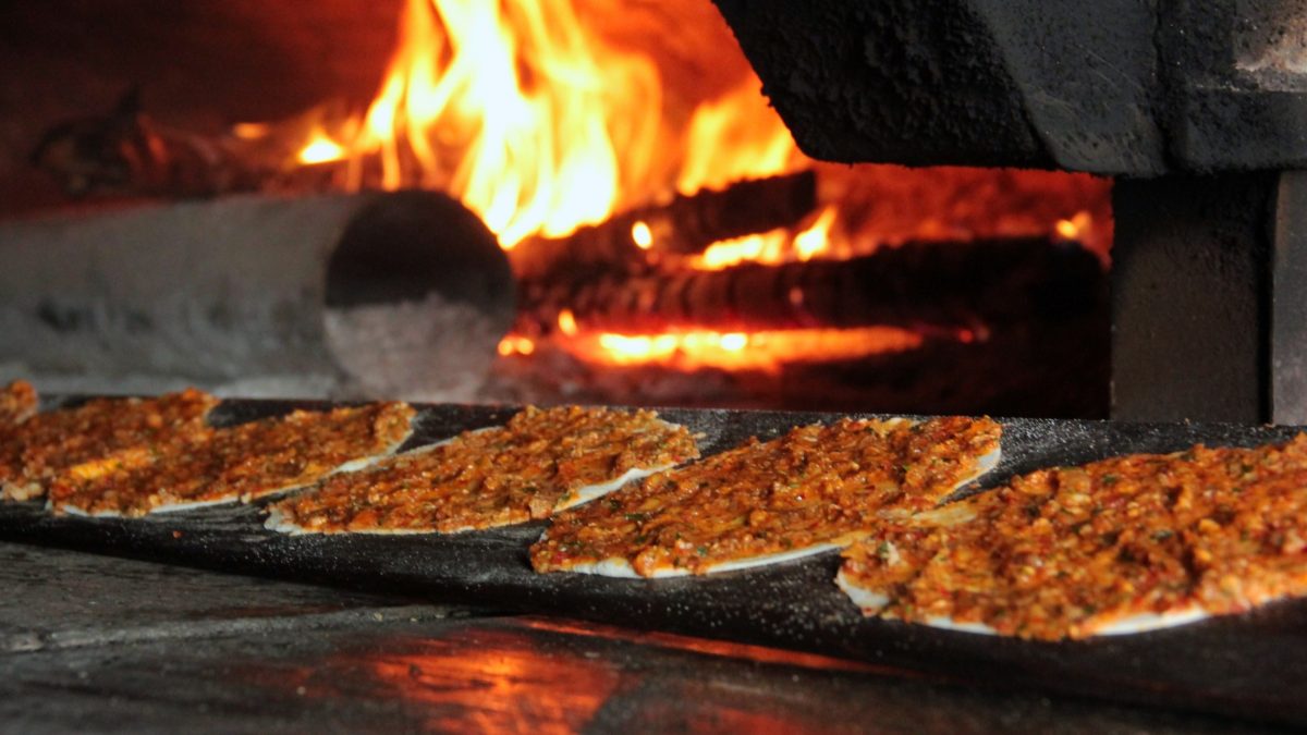 Frontal: Vier Fladen mit Hackfleisch werden in einen offenen Ofen, in dem ein Feuer brennt, geschoben. Der Fladen wurde nach Türkische Hackfleisch-Rezept zubereitet,