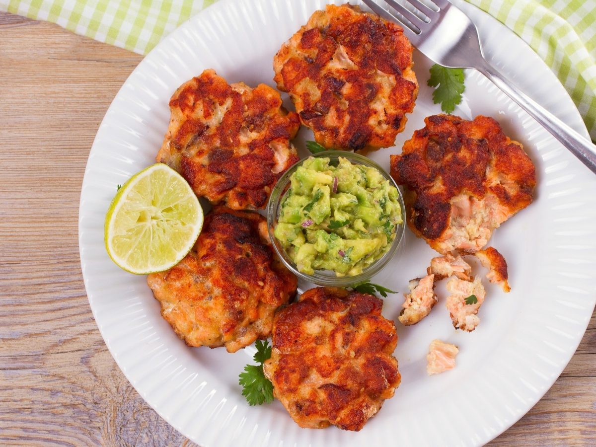 Draufsicht: Selbst gemachte Lachs-Patties auf einem weißen Teller. In der Mitte steht ein Glasschälchen mit Avocado-Creme. Auf dem teller liegt eine Gabel. Und neben dem Teller ein hellgrün und weiß kariertes Handtuch.