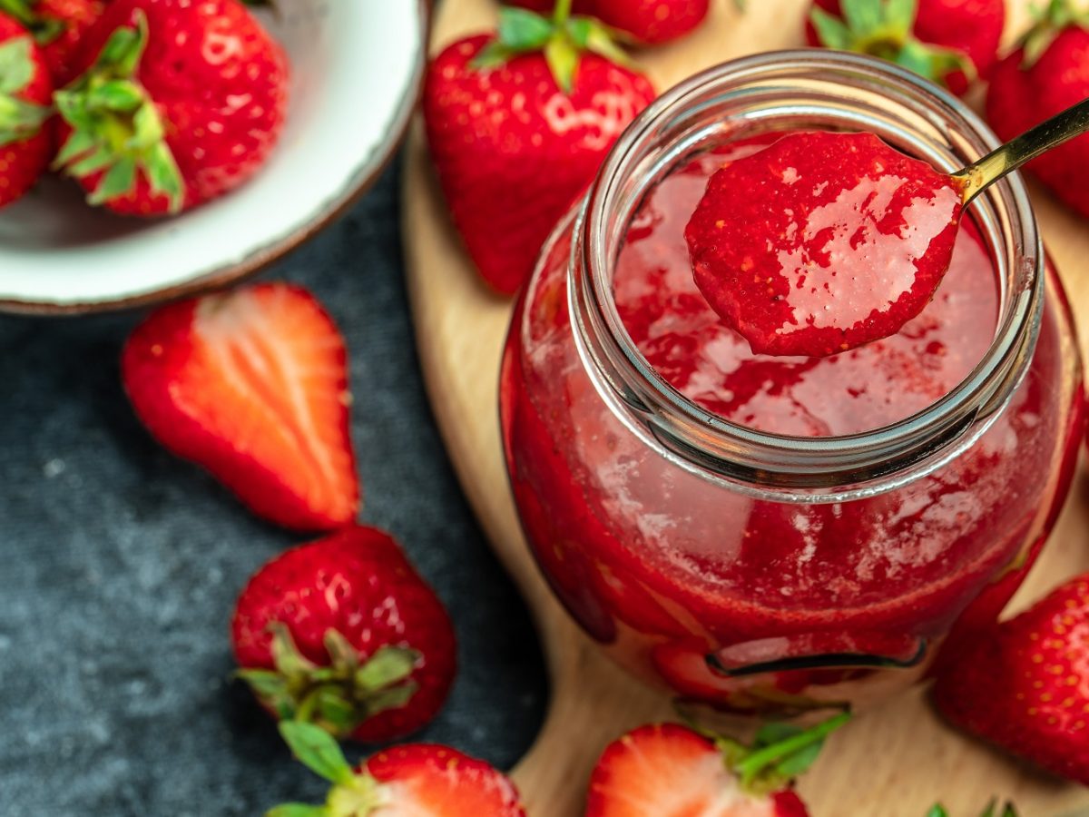 Ein Löffel hebt Erdbeermarmelade aus einem Glas, umgeben von reifen Erdbeeren auf einem dunklen Untergrund.