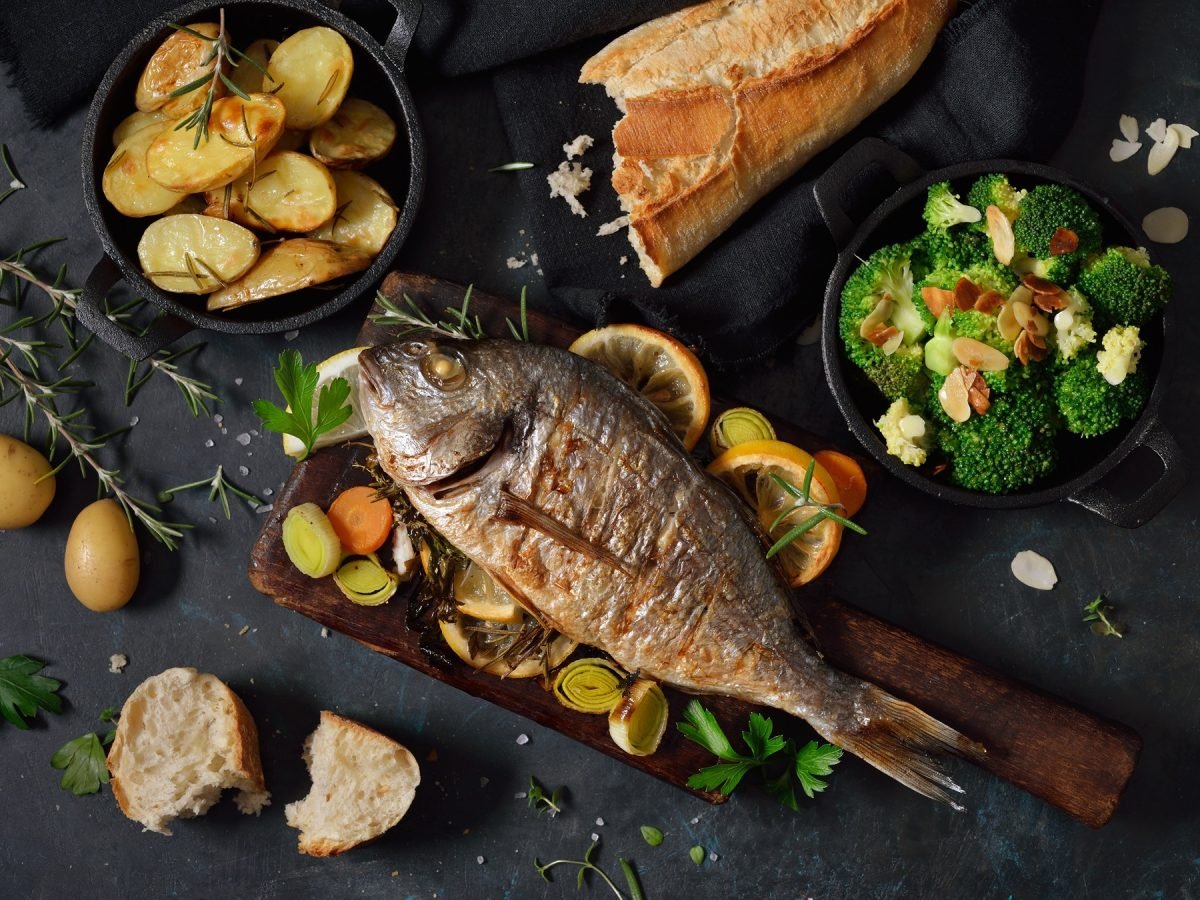 Draufsicht auf ein Fisch-gericht mit gegrillter Dorade und knusprigen Kartoffeln vor einem dunklen Untergrund.