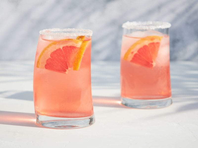 Orange-pinkfarbener Cocktail garniert mit Grapefruit in zwei gebogenen Gläsern.