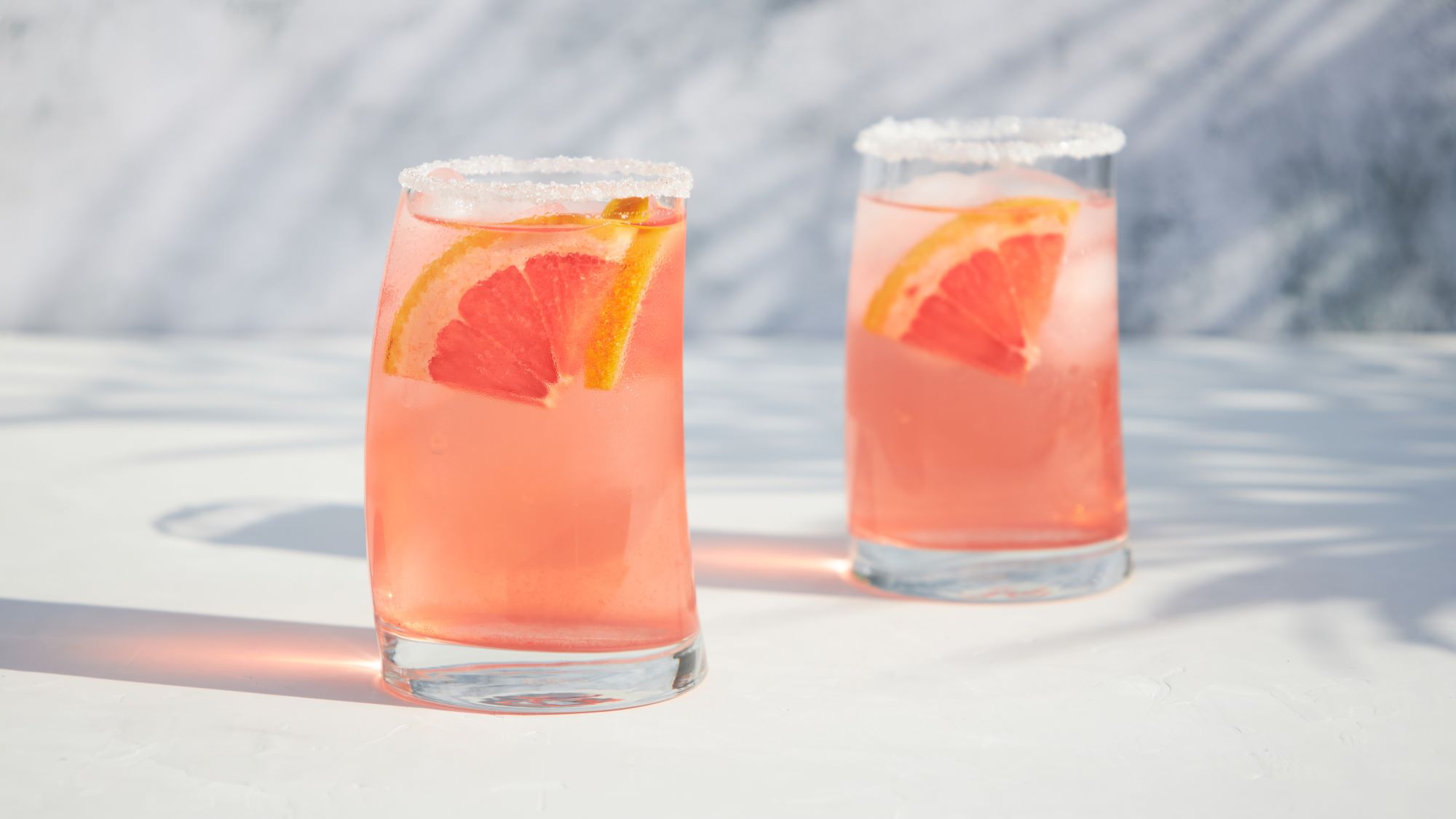 Orange-pinkfarbener Cocktail garniert mit Grapefruit in zwei gebogenen Gläsern.