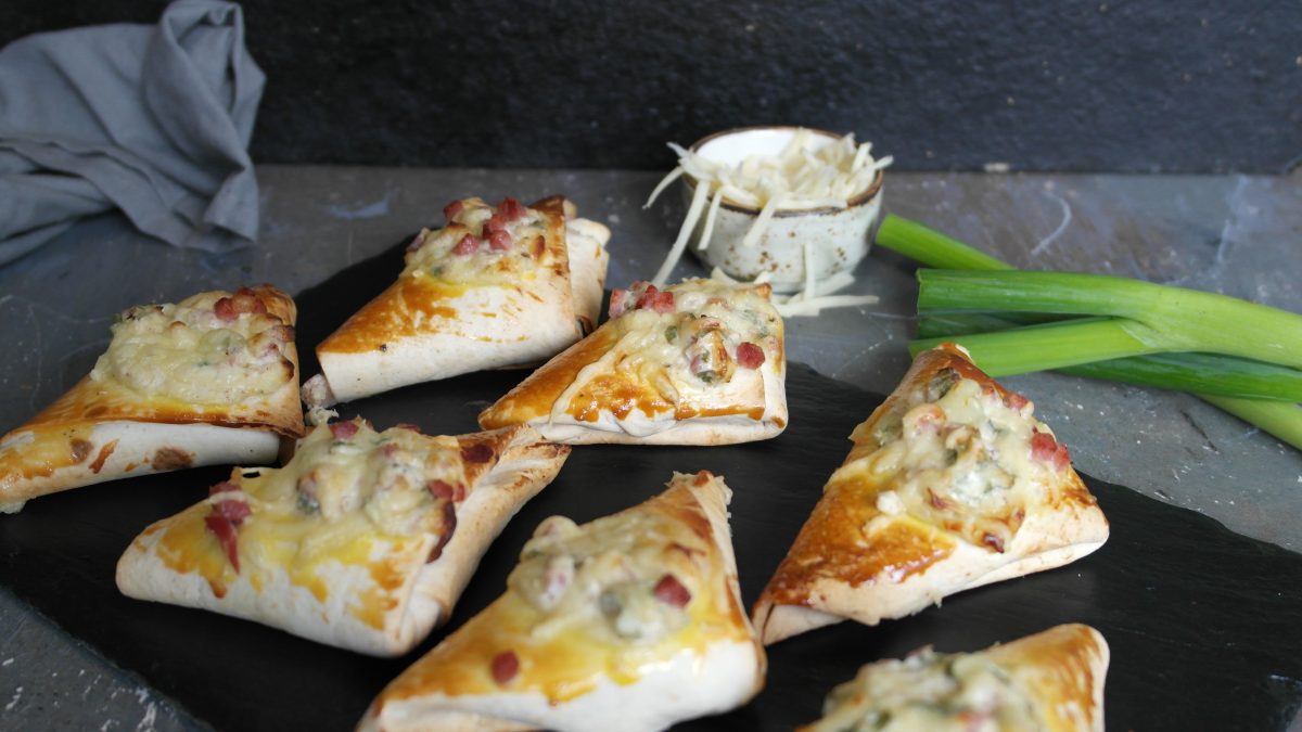 Eine schwarze Schiefertafel mit den Flammkuchen-Taschen auf einem grauen Steintisch. Dahinter Reibekäse und Frühlingslauch vor schwarzem Hintergrund. Alles von leicht schräg oben fotografiert.