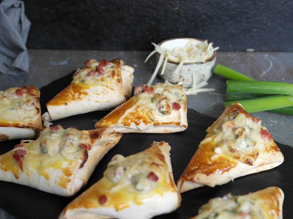 Eine schwarze Schiefertafel mit den Flammkuchen-Taschen auf einem grauen Steintisch. Dahinter Reibekäse und Frühlingslauch vor schwarzem Hintergrund. Alles von leicht schräg oben fotografiert.