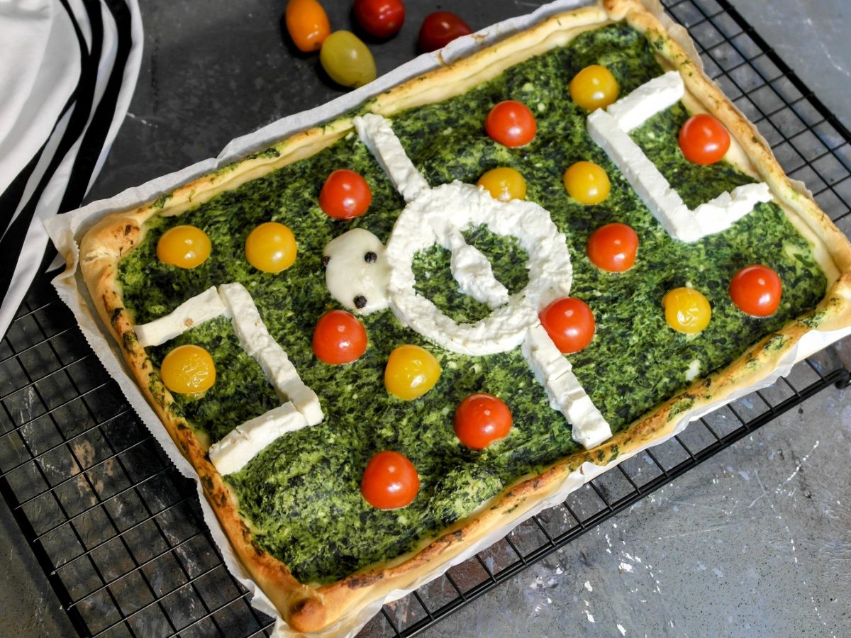 Die Fussball-Pizza auf einem schwarzen Metallgitter und grauem Steintisch. Daneben ein paar bunte Kirschtomaten und eine weiß-schwarze Fußball-Shorts. Alles von leicht schräg oben fotografiert.