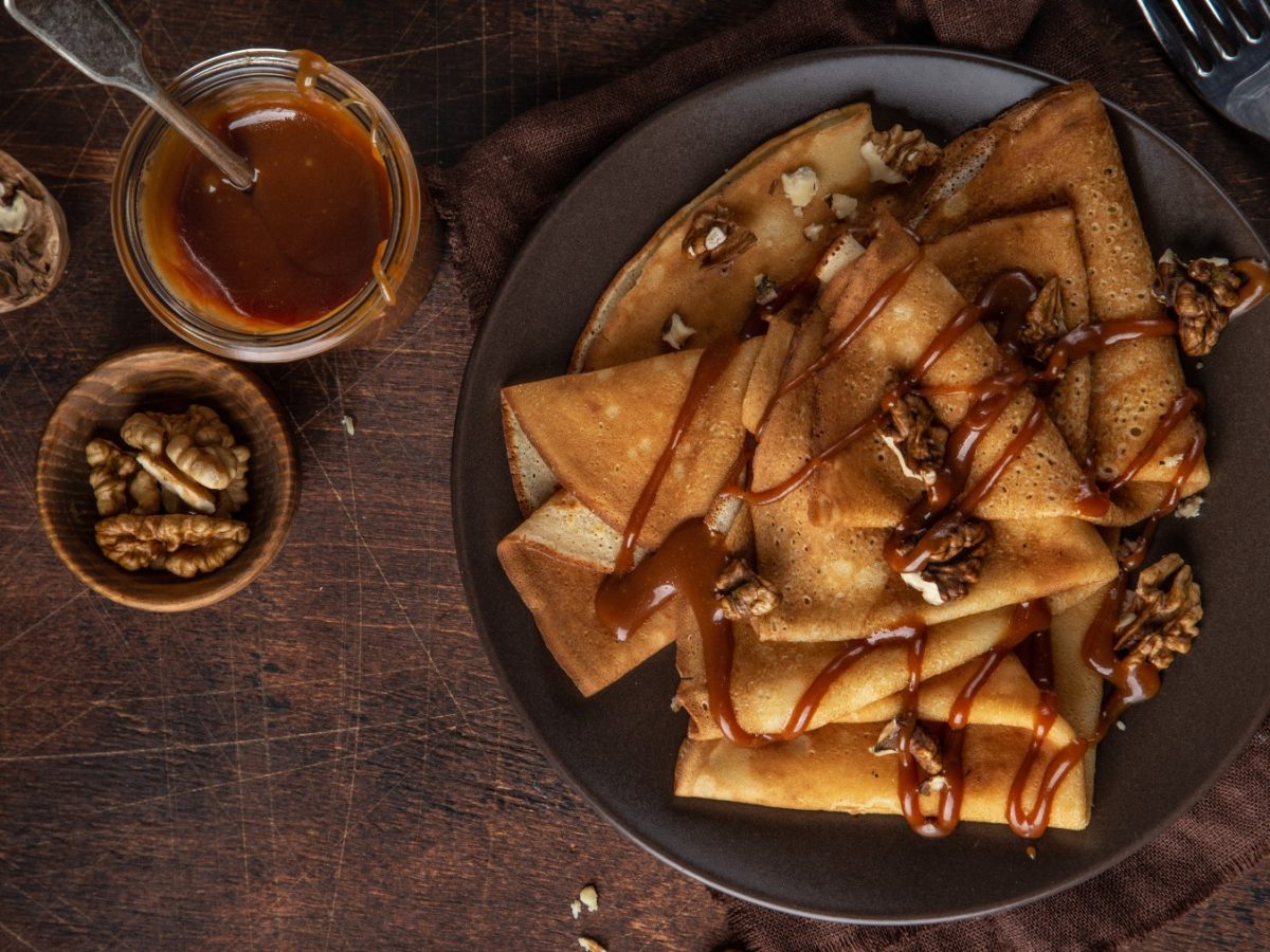 Glutenfreie Crêpes auf einem schwarzen Teller auf braunem Hintergrund serviert mit Walnüssen und Karamellsauce.