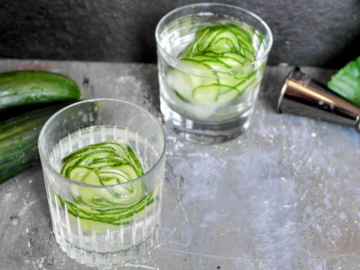 Zwei Gläser mit den Gurken-Eiswürfeln auf einem Steintisch mit schwarzem Hintergrund.