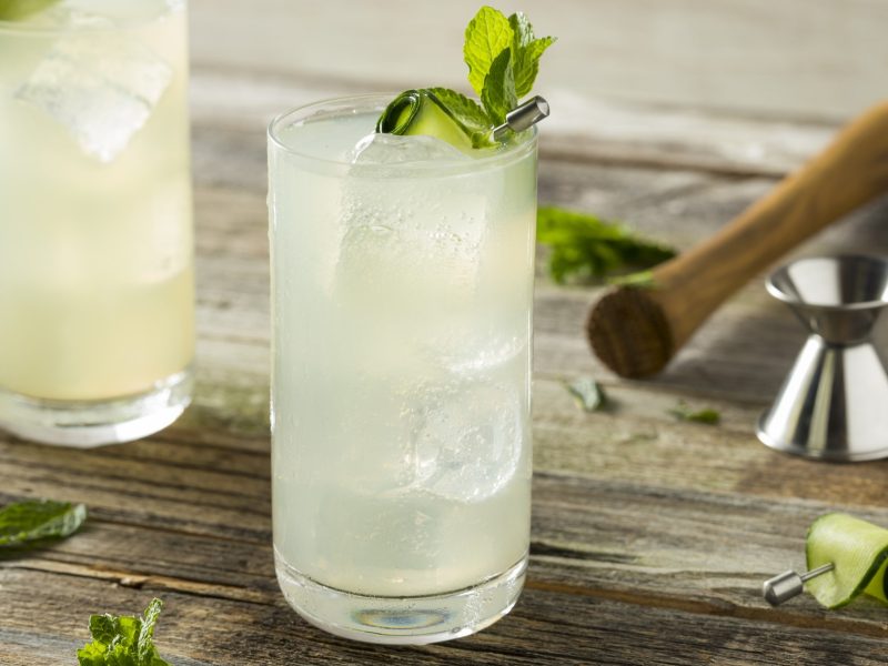 Zwei Gläser mit dem Gurken-Spritz auf einem rustikalen Holztisch mit Minze, Gurke und Barzubehör. Alles von der Seite fotografiert.