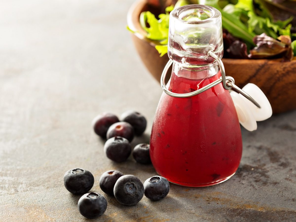 Heidelbeer-Vinaigrette in einer verschließbaren Flaschen, daneben Heidelbeeren und im Hintergrund eine Schüssel Salat.