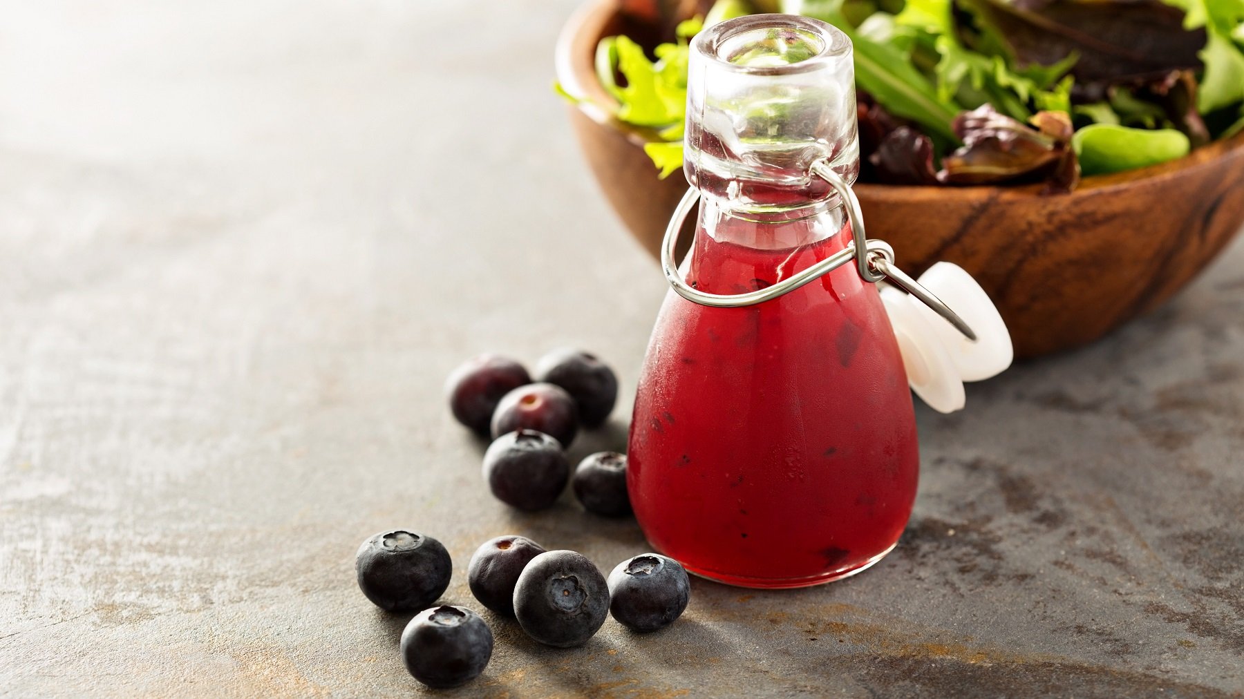 Heidelbeer-Vinaigrette in einer verschließbaren Flaschen, daneben Heidelbeeren und im Hintergrund eine Schüssel Salat.