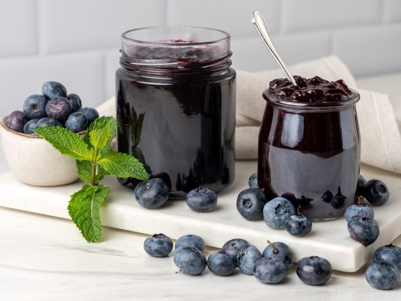 Gläser mit dunkler Heidelbeermarmelade und frische Blaubeeren auf weißem Brett in heller Küche arrangiert.