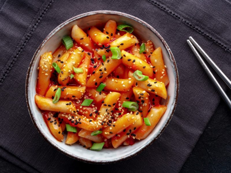 Honig-Butter-Tteokbokki in einer Schüssel auf schwarzem Hintergrund. Rechts auf dem Bild Stäbchen.