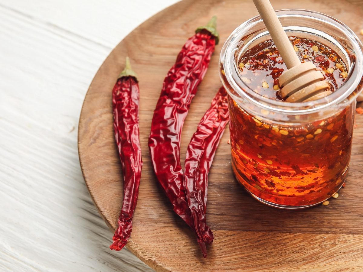 Hot Honey von schräg oben fotografiert in einem Glas mit Honiglöffel, daneben getrocknete Chilischoten.