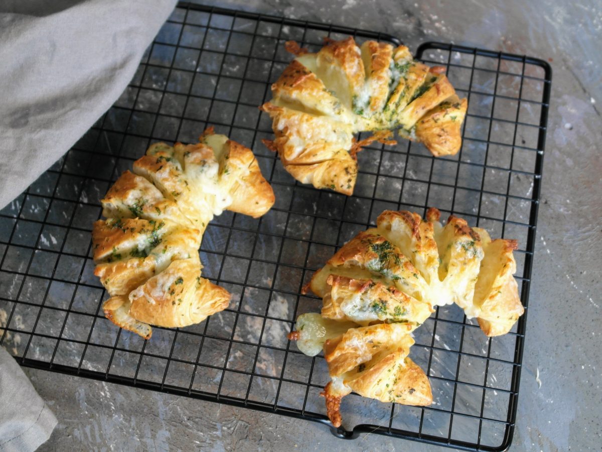 Drei Käse-Kräuter-Croissants auf einem schwarzen Gitter und grauem Untergrund direkt von oben fotografiert.