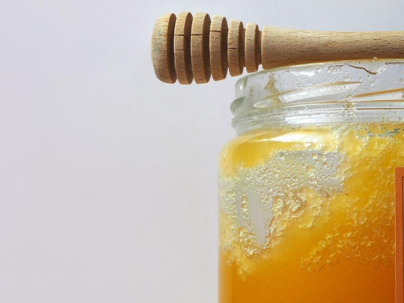 Ein Glas mit dem Honig und einem Holzlöffel auf vor grauem Hintergrund sehr nah fotografiert.